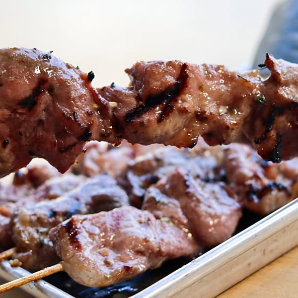 Close-up of a grilled Thai pork skewer with char marks, glistening with sauce, above a tray of more skewers.