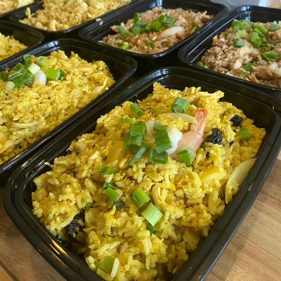 Multiple black containers filled with Thai fried rice, garnished with green onions, shrimp, and raisins.