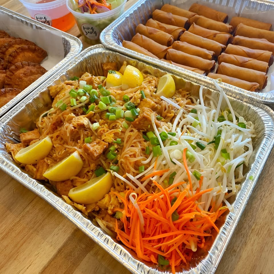 Thai catering spread with Pad Thai, spring rolls, dumplings, and sauces in foil containers.